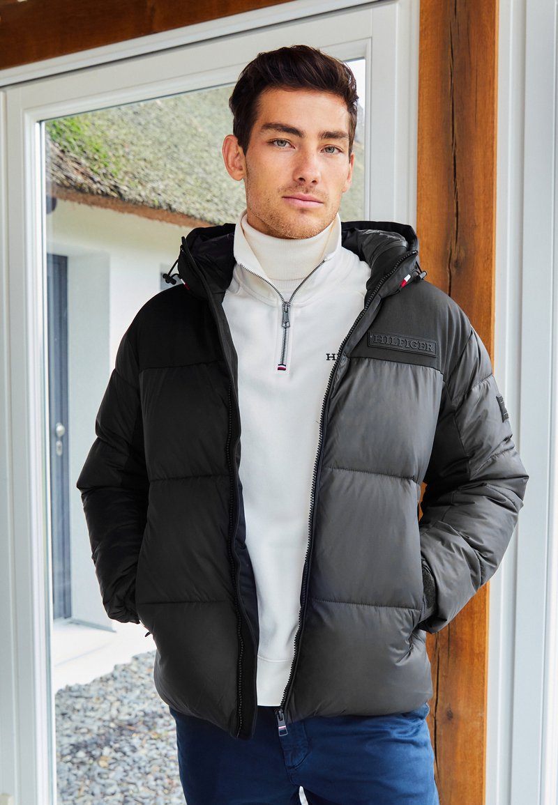 Č Black padded jacket with a hood, zip closure, and logo on the chest, paired with a white high-neck sweater and dark blue pants.