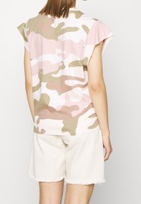 Tričko s krátkým rukávem v růžovém maskáčovém vzoru vyrobené z měkké tkaniny, s volným střihem a mírně nařasenými rukávy, kombinované s bílými šortkami.