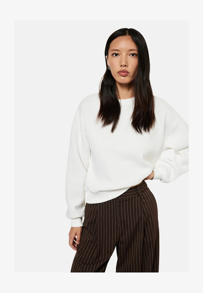 Woman with long dark hair wears white oversized sweater and high-waisted brown pinstripe pants, standing with one hand on hip.