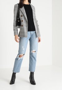 Grau karierter doppelt-mantelloser Blazer mit braunen Knöpfen, kombiniert mit einem schwarzen Oberteil. Hellblaue, ausgewaschene Jeans und schwarze Stiefeletten.