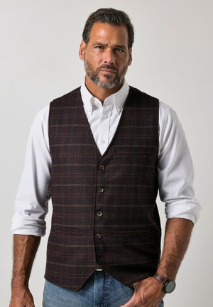 Man met baard die een wit overhemd met opgerolde mouwen, een donker geruit gilet, blauwe spijkerbroek en een bruin leren horloge draagt, staat voor een effen achtergrond.