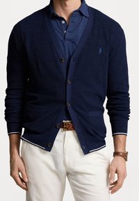 Mörkblå stickad cardigan med v-ringad hals, knappstängning, två framfickor och kontrasterande vita detaljer på ärmsluten. Bärs över en mörkblå polo.