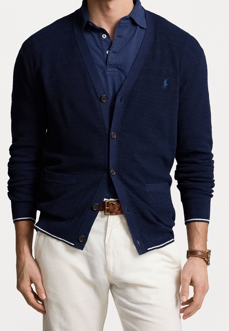 Mörkblå stickad cardigan med v-ringad hals, knappstängning, två framfickor och kontrasterande vita detaljer på ärmsluten. Bärs över en mörkblå polo.