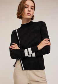 Schwarzer gerippter Pullover mit hohem Kragen, der weiße Streifenakzente an den Ärmeln und einen seitlichen Schlitz für zusätzlichen Detailreichtum aufweist.