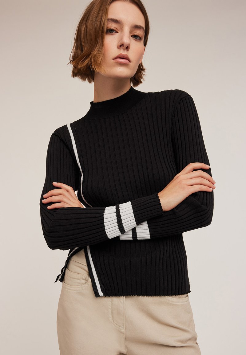 Schwarzer gerippter Pullover mit hohem Kragen, der weiße Streifenakzente an den Ärmeln und einen seitlichen Schlitz für zusätzlichen Detailreichtum aufweist.