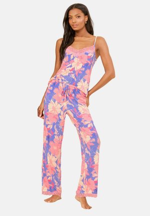 Ung kvinde iført lilla og pink blomstret camisole med blonder og matchende pyjamasbukser med snøre, stående barfodet.