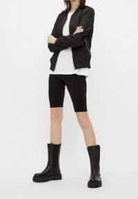 Persona de pie con los brazos cruzados, vistiendo una chaqueta negra con cremallera, una camiseta blanca, pantalones cortos negros y botas de plataforma altas negras sobre un fondo blanco.