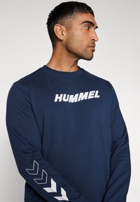 Mørkeblå langermet skjorte lavet af blødt materiale har et hvidt "HUMMEL" logo på brystet og hvide chevron-detaljer på ærmerne.