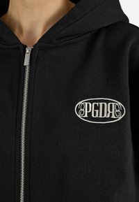 Felpa nera con zip realizzata in tessuto morbido, caratterizzata da un logo ricamato bianco sul lato sinistro del petto. Include un cappuccio con coulisse e una zip argentata.