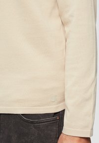 Langarm, licht beige shirt van textuurstof met een klein logo aan de zoom. Gepaard met donkergrijze broek met een zak.