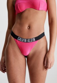 Růžové žebrované tanga s černým pasem s logem "CALVIN KLEIN". Elastický materiál s těsným střihem pro pohodlí a oporu.
