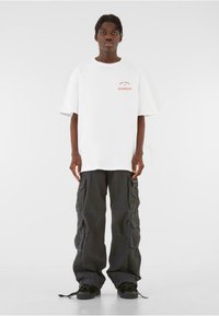 White oversized t-shirt featuring red text. Paired with loose-fitting dark gray cargo pants with multiple pockets. Black sneakers.