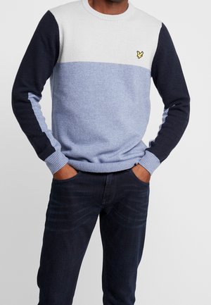 Mand iført en farvebloktrøje med hvide, lyseblå og mørkeblå sektioner og mørke jeans, hænderne i lommerne, stående mod en ensfarvet baggrund.