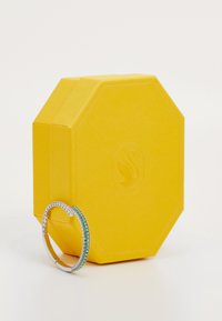 Boîte hexagonale jaune avec une surface texturée à côté d'un bracelet en argent orné de pierres précieuses claires et vertes le long de la bande.