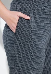 Handen är inskjuten i framfickan på mörkgrå byxor med en liten diamantmönster av tygtextur.