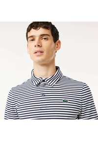 Marinblå och vit randig pikétröja med krage, försedd med tre knappar och en grön krokodillogo på vänster sida. Tillverkad av bomull.