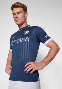 Camiseta deportiva azul marino con rayas verticales, cuello y acentos en las mangas blancos. Presenta un logo en el pecho y está hecha de un tejido suave y ligero.