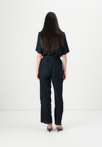 Jumpsuit blu navy con maniche corte, vita arricciata e pantaloni a gamba larga. Completato da un orlo leggermente sfilacciato e indossato con sandali con tacco.