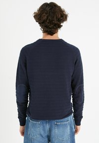 Suéter texturizado de color azul marino con mangas largas y puños acanalados. Presenta parches en los codos; la vista posterior muestra un diseño acanalado y un cuello redondo.