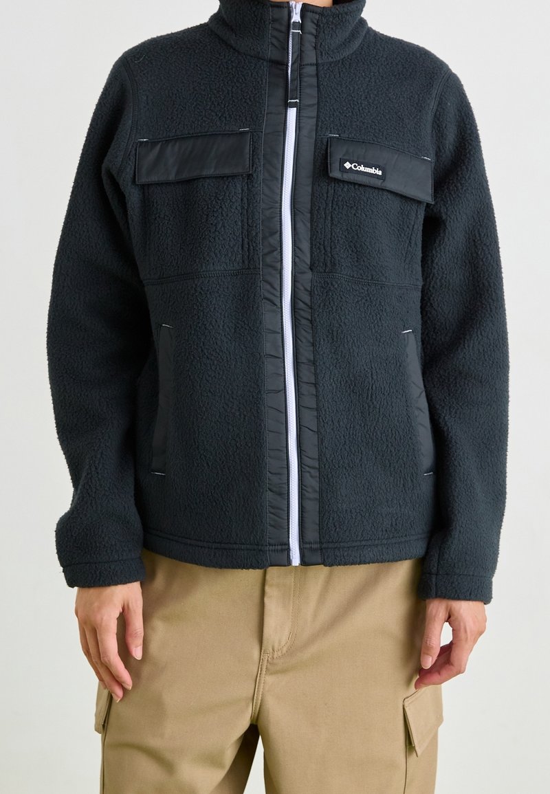 Personne portant une veste polaire noire Columbia avec une fermeture éclair frontale et deux poches poitrine, associée à un pantalon cargo beige.