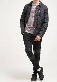 Chaqueta negra acolchada sobre un suéter de punto con patrones de diseños geométricos. Pantalones negros con detalles de panel de cuero. Zapatillas negras y calcetines blancos.