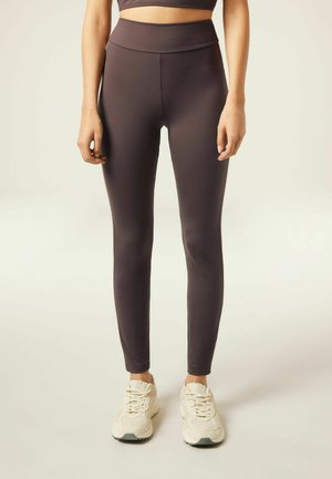 Hochgeschnittene, schmal geschnittene Leggings aus dehnbarem dunkelbraunem Material, kombiniert mit hellen Sportschuhen. Glatte Textur, keine sichtbaren Muster.