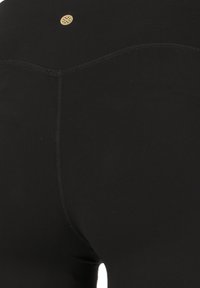 Zwarte leggings van rekbaar materiaal, met een naadloos ontwerp, gebogen achterste stiksels en een klein gouden logo detail nabij de tailleband.