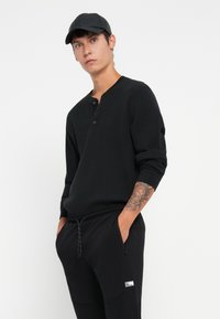 Crna rebrasta majica s dugim rukavima i dugmadi, u kombinaciji s crnim tapered hlačama s elastičnom waistbandom i logotipom.