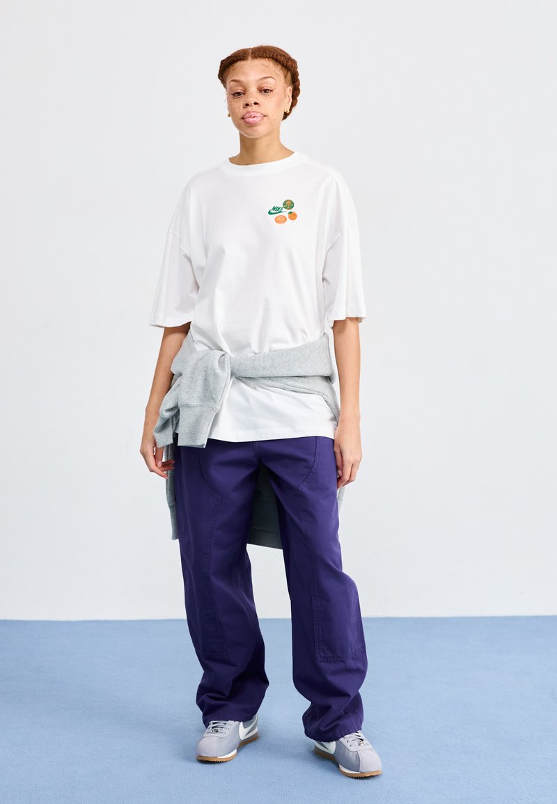 Camiseta blanca de gran tamaño con gráficos en verde y naranja, suéter gris atado a la cintura, pantalones morados y zapatillas grises con suela de goma.