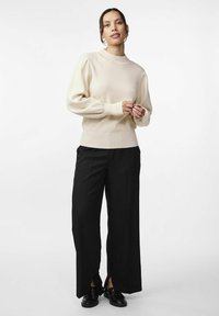 Beiger gestrickter Pullover mit hohem Kragen und puffigen Ärmeln, kombiniert mit schwarzen weit geschnittenen Hosen und schwarzen Schuhen. Glatte Textur, moderne Passform.