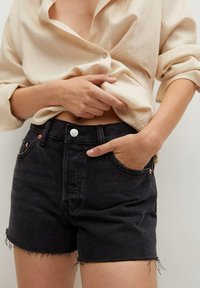 Pantalones cortos de mezclilla negros con dobladillo deshilachado, diseño de cintura alta y sutiles acentos en cobre. Combinados con una camisa abotonada de textura ligera en color beige.