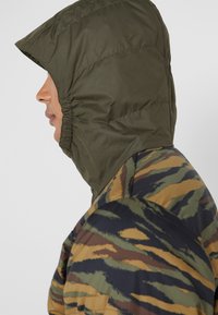 Vue de profil d'une personne portant une capuche vert olive et une doudoune avec un motif camouflage vert, marron et noir.
