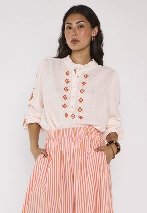 Donna che indossa una camicetta bianca con ricamo floreale arancione e una gonna a righe rosa-arancione, mani nelle tasche, in piedi contro uno sfondo semplice.