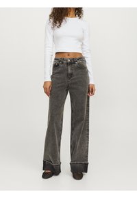Frau in einem weißen langärmeligen verkürzten Oberteil, hochtaillierten, weit geschnittenen, verwaschenen schwarzen Jeans mit umgeschlagenen Säumen und schwarzen Netzschuhen.