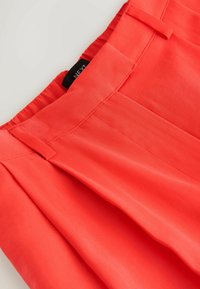 Gros plan sur la ceinture d'un pantalon orange corail vif avec des passants de ceinture, des plis et une étiquette de vêtement noire partiellement visible à l'intérieur.