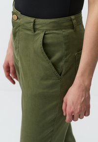 Pantaloni in cotone verde oliva con vita alta, dotati di una tasca laterale e chiusura con bottone. Il tessuto ha una consistenza liscia.