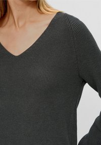 Frau, die einen dunkelgrauen, langärmeligen Strickpullover mit V-Ausschnitt trägt, von Hals bis Taille vor einem einfarbigen Hintergrund gezeigt.