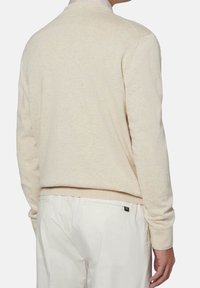Mann in einer cremefarbenen, langärmligen Sweater und weißen Hosen, von hinten gegen einen schlichten hellen Hintergrund gezeigt.
