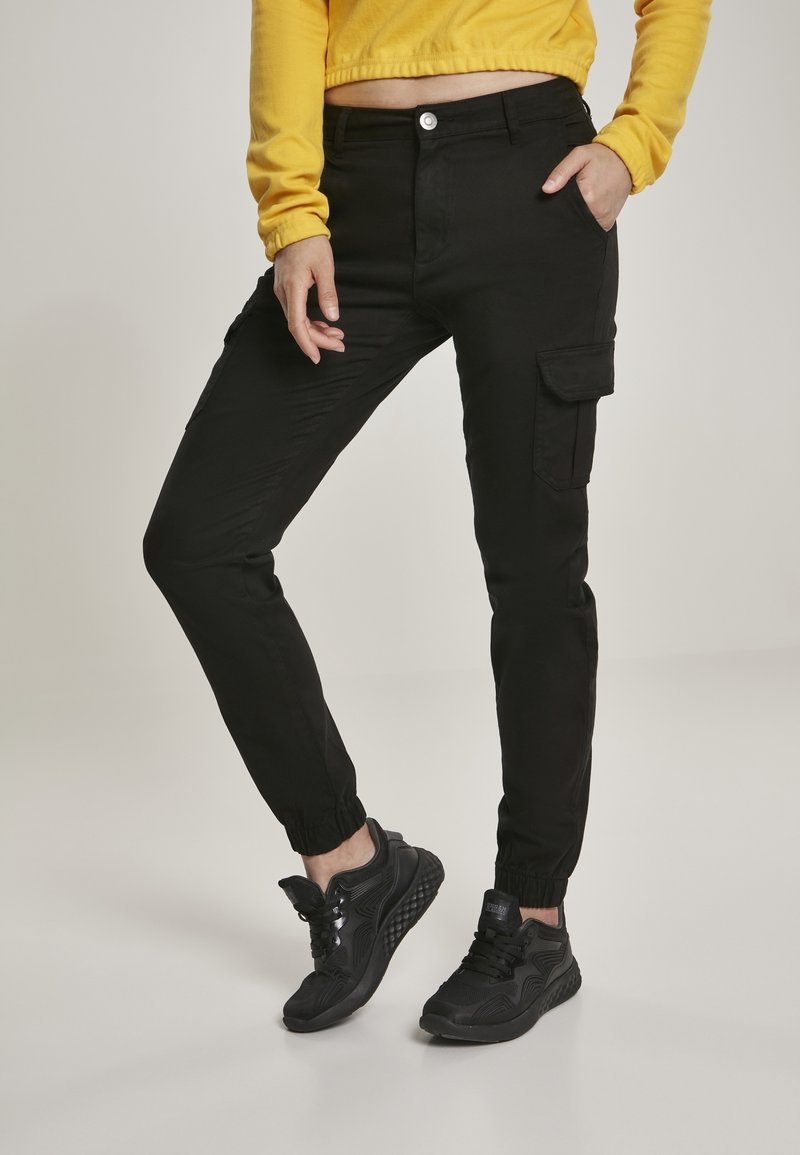 Personne portant un pantalon cargo jogger noir et des baskets noires, posant avec une main dans la poche et un sweat jaune court.