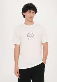 Hvit bomulls t-skjorte med korte ermer, rund hals og en svart sirkulær logo med "A/X" i midten. Casual passform.