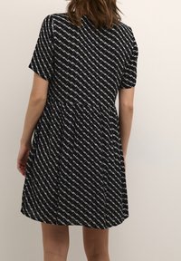 Schwarzes Kleid mit kurzen Ärmeln und weißem geometrischem Muster, ausgestelltem Rock und weicher Textur. Rückansicht zeigt eine lockere Passform.