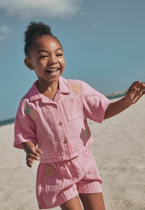Lächelndes Kind mit lockigem Haar in pinken, passenden Shorts und Shirt mit gestickten Motiven, das an einem sonnigen Sandstrand spielt.
