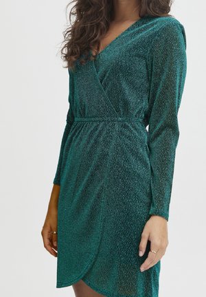 Mujer vestida con un vestido cruzado verde brillante de manga larga, con cintura entallada, posando frente a un fondo claro y liso.