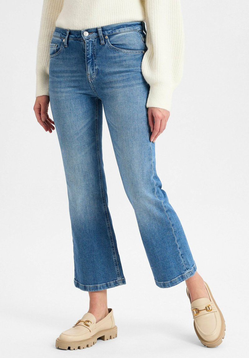 Vrouw in lichtblauwe, cropped flare jeans, crèmekleurige ribgebreide trui en beige instappers met gouden gesp-details op een witte achtergrond.