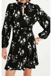 Zwarte bloemenjurk met pofmouwen. Bevat gele en witte bloemenpatronen, een sleutelgaatjeshalslijn en een getailleerde taille.