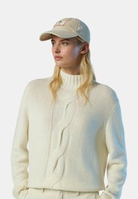 Maglione bianco lavorato a maglia con collo alto e motivo a treccia. Abbinato a un cappellino beige con patch logo. Texture morbida, vestibilità comoda.