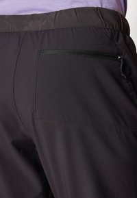 Zwarte broek van een gladde, lichte stof met een elastische tailleband. Een ritszak is zichtbaar aan de achterkant.