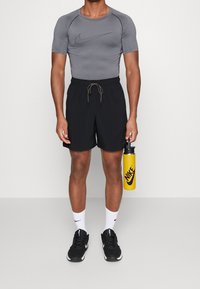 Chemise athlétique grise, shorts noirs avec cordon de serrage, baskets noires, chaussettes blanches, tenant une bouteille d'eau jaune avec logo Nike.