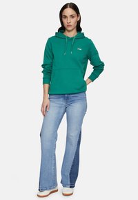 Sudadera verde de Fila con cordón, bolsillo frontal y puños acanalados. Combinada con jeans de campana de color azul claro que cuentan con un panel lateral de mezclilla oscura.