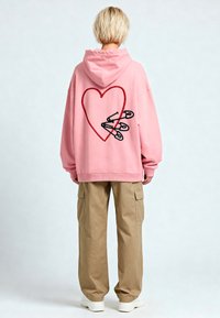 Felpa oversize rosa con un design di cuore rosso e spille da balia nere ricamate sul retro. Materiale in cotone, con cappuccio regolabile.